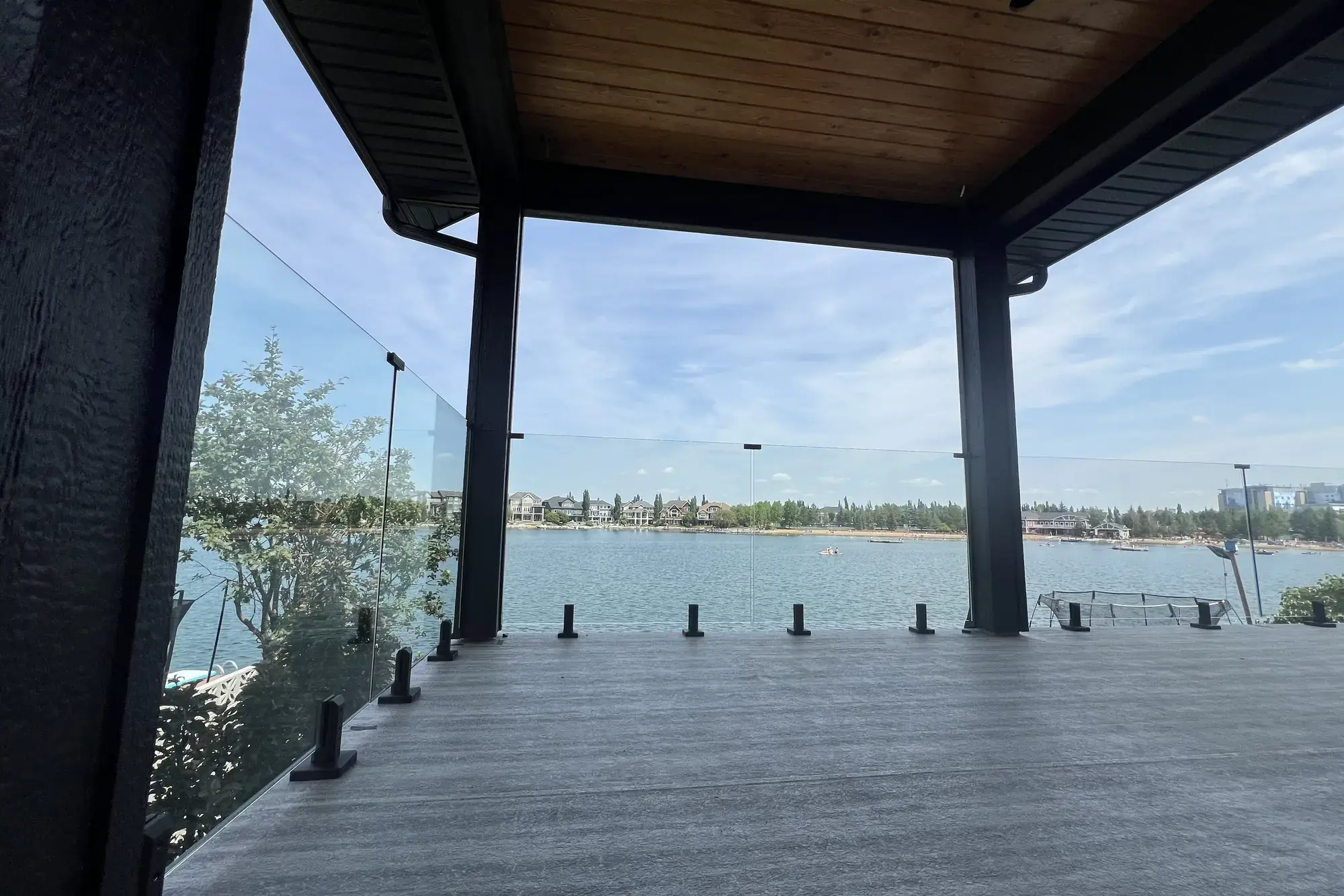 A covered deck on a new lakefront home, woodgrain vinyl surface under a timber ceiling and glass railing out over the water